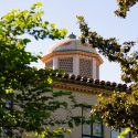 A decorative dome with floral patterns rises above a tiled roof, framed by lush green foliage against a clear blue sky.