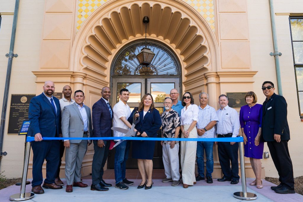 A group of people stands together in front of a building, holding a large ceremonial scissors to cut a blue ribbon during an official opening.