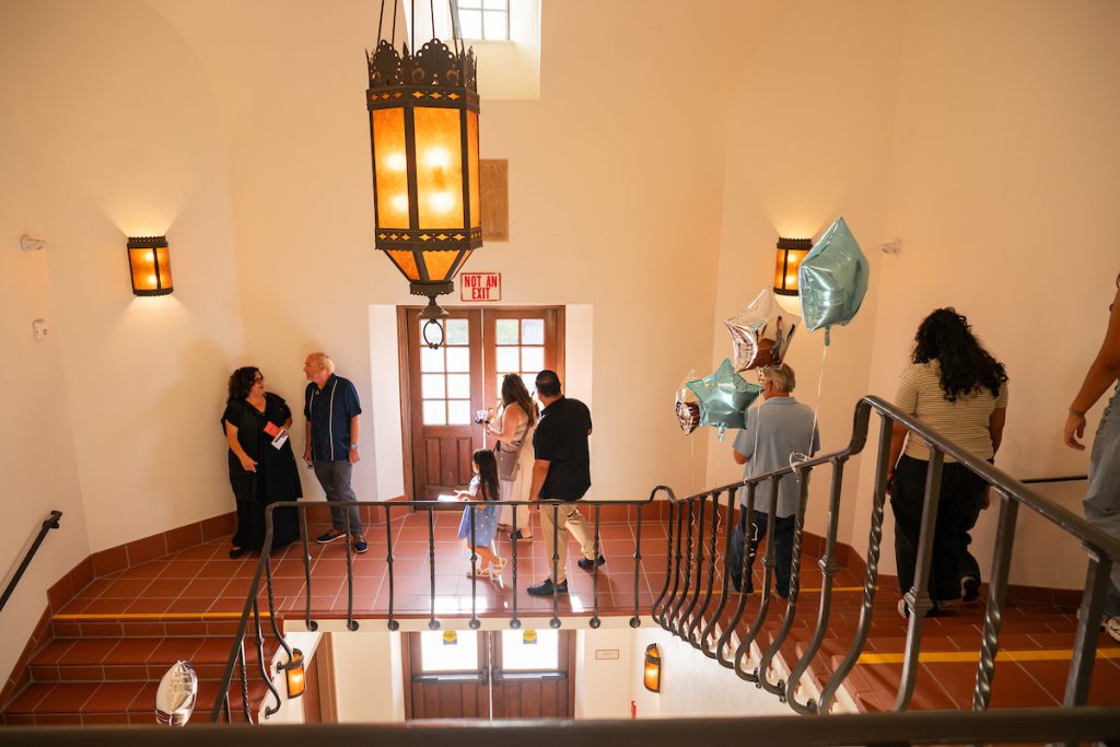 People gather in a warmly lit hallway with tiled floors and ornate lighting, some holding balloons as they converse near a doorway marked “Not an Exit.”
