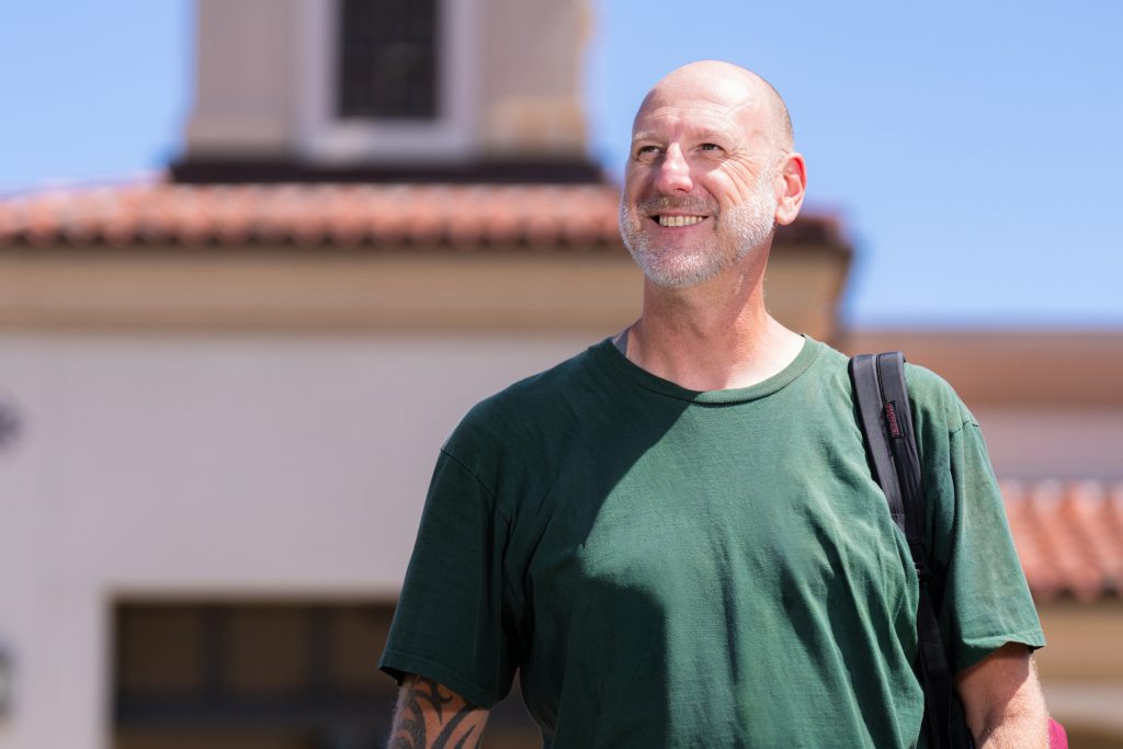 A bald individual with a beard smiles while standing outdoors in front of a building with a tiled roof, wearing a green t-shirt and carrying a backpack.